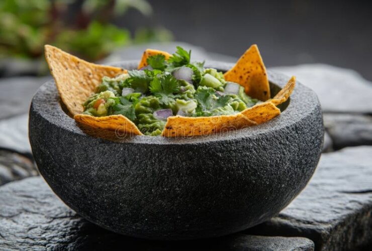 A rustic stone bowl filled with chunky restaurant style guacamole garnished with fresh cilantro leaves, sliced jalapeño, and a wedge of lime, surrounded by tortilla chips on a dark wood surface