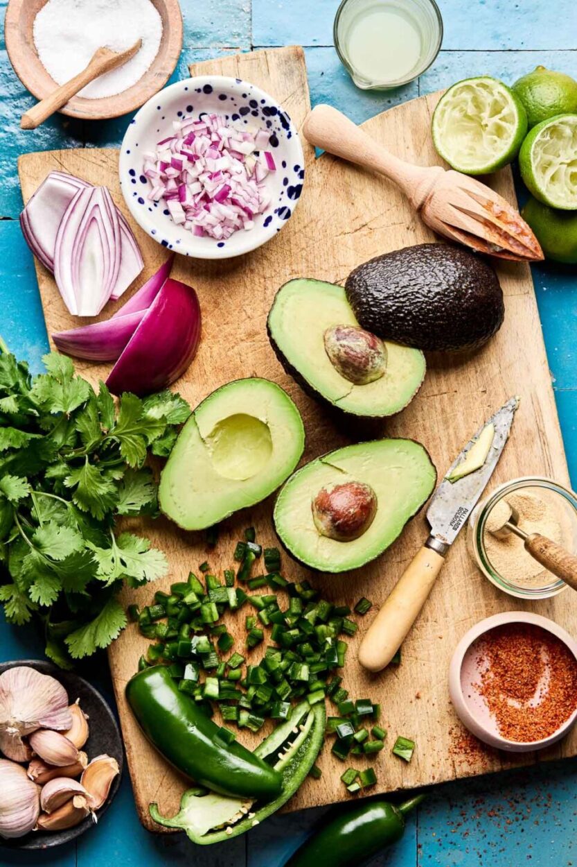 Overhead flat-lay of fresh guacamole ingredients on a white marble surface — ripe Hass avocados halved and pitted, quartered limes, diced white onion, sliced jalapeño, fresh cilantro sprigs, coarse salt, and a small garlic clove