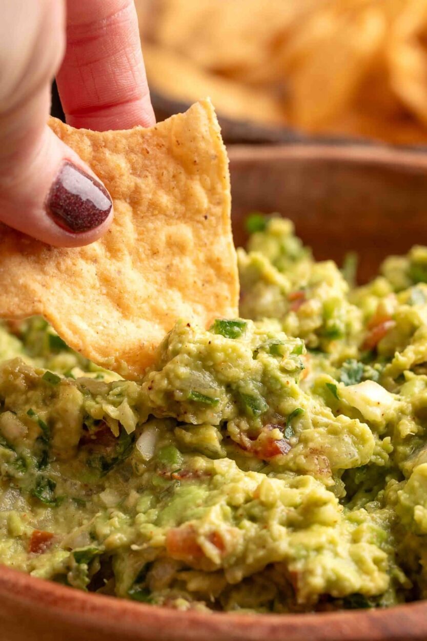 Close-up of restaurant style guacamole being scooped with a tortilla chip, showing the chunky avocado texture, bright green color, and visible pieces of cilantro and jalapeño