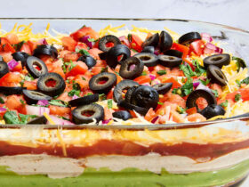 Seven layer dip in a clear glass dish showing all colorful layers — refried beans, guacamole, sour cream, cheese, salsa, black olives, and green onions — served with tortilla chips on a party platter