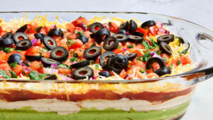 Seven layer dip in a clear glass dish showing all colorful layers — refried beans, guacamole, sour cream, cheese, salsa, black olives, and green onions — served with tortilla chips on a party platter
