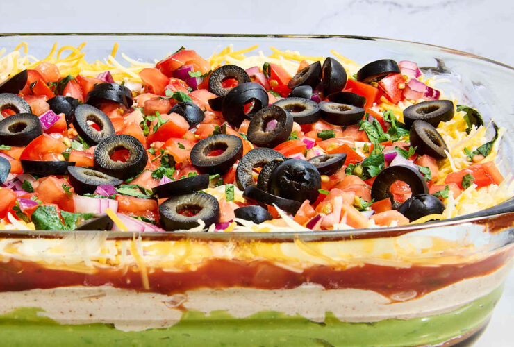 Seven layer dip in a clear glass dish showing all colorful layers — refried beans, guacamole, sour cream, cheese, salsa, black olives, and green onions — served with tortilla chips on a party platter