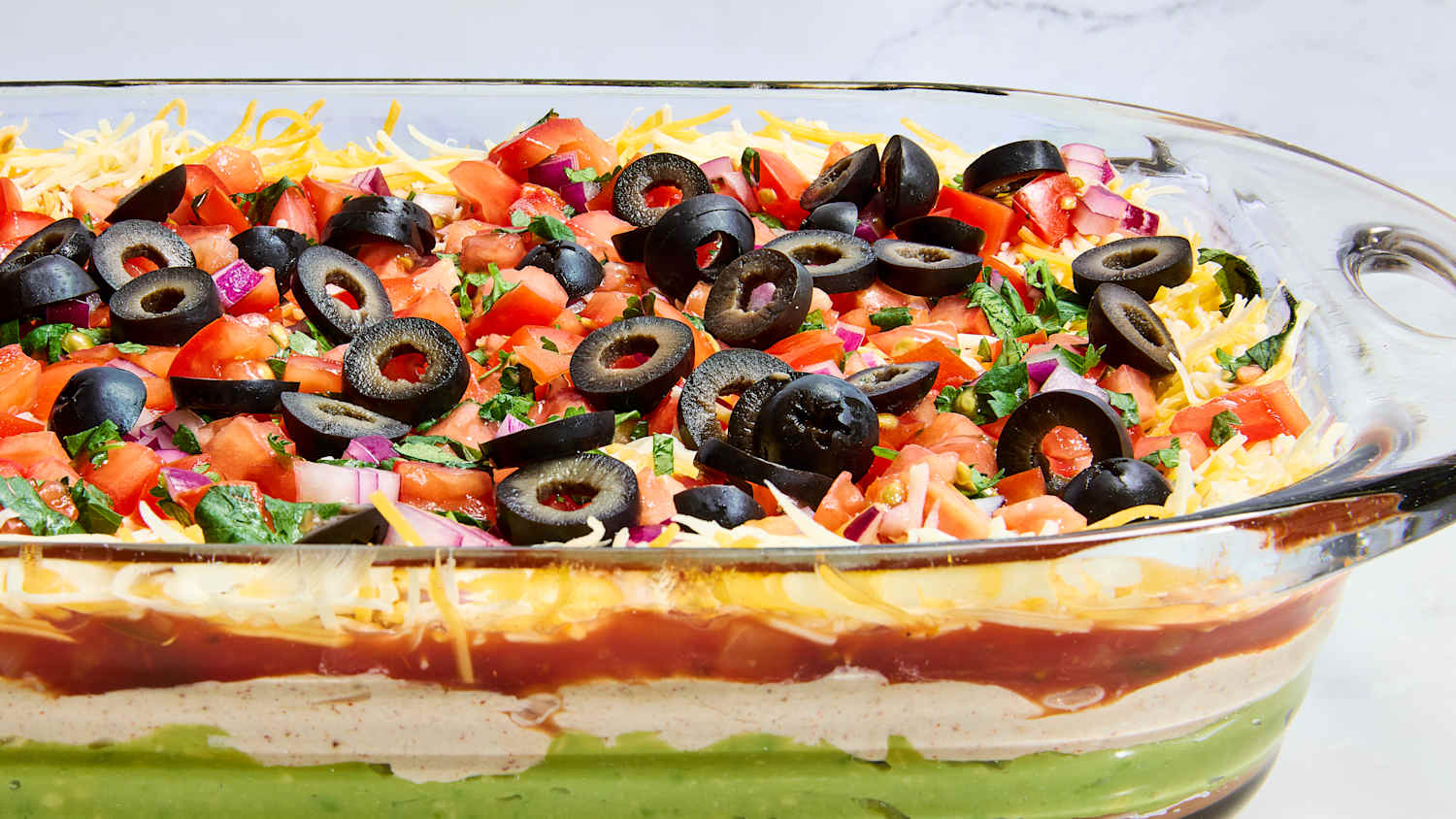 Seven layer dip in a clear glass dish showing all colorful layers — refried beans, guacamole, sour cream, cheese, salsa, black olives, and green onions — served with tortilla chips on a party platter