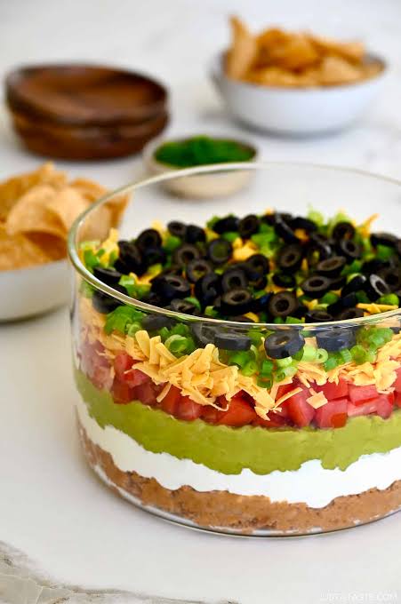 Overhead flat-lay of seven layer dip ingredients — cans of refried beans, ripe avocados, sour cream, shredded cheese, salsa jar, black olives, and green onions arranged on a wooden board