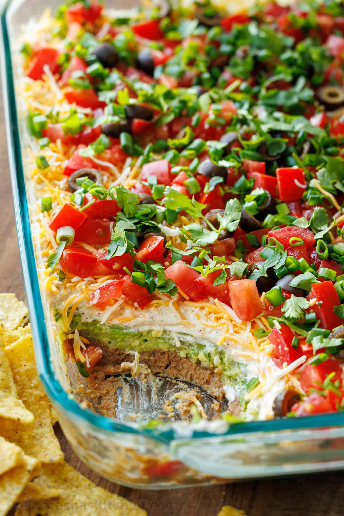 Close-up of a tortilla chip being scooped through all seven layers of the dip — showing beans, guacamole, sour cream, cheese, salsa, and toppings on a single chip