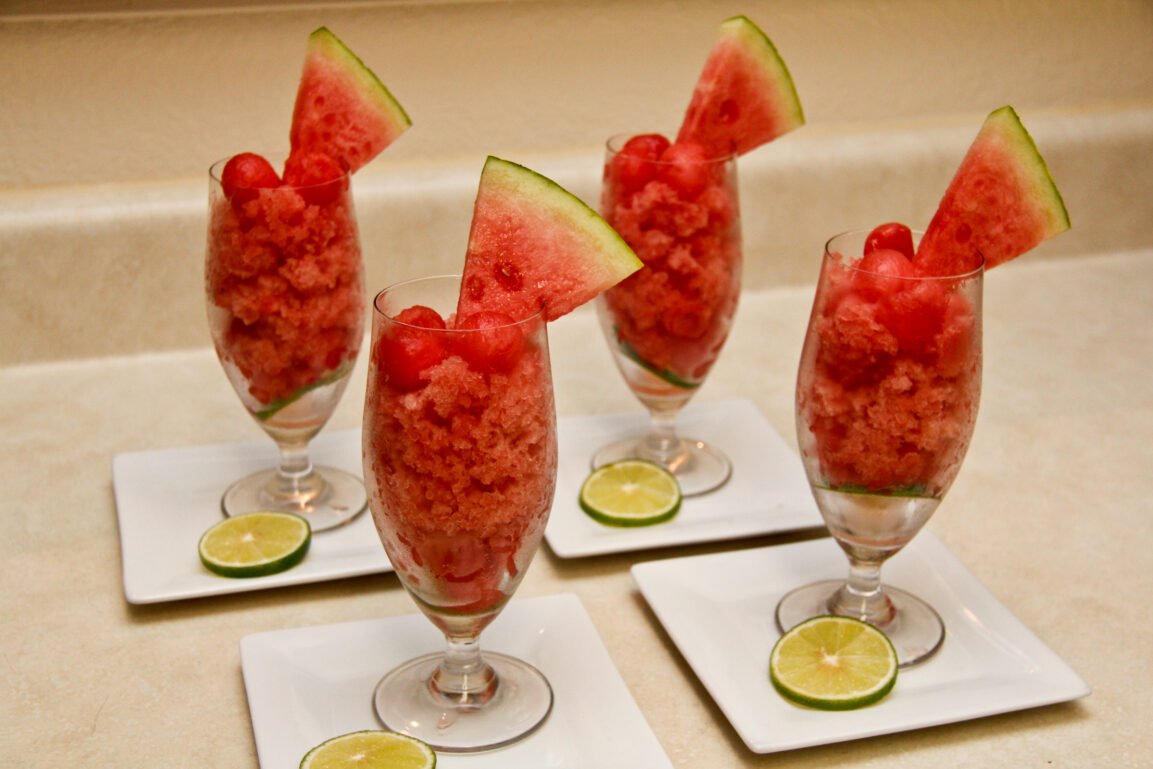 Watermelon lime granita served in chilled glasses with fresh lime wedges and mint leaves on a rustic wooden table in bright summer light