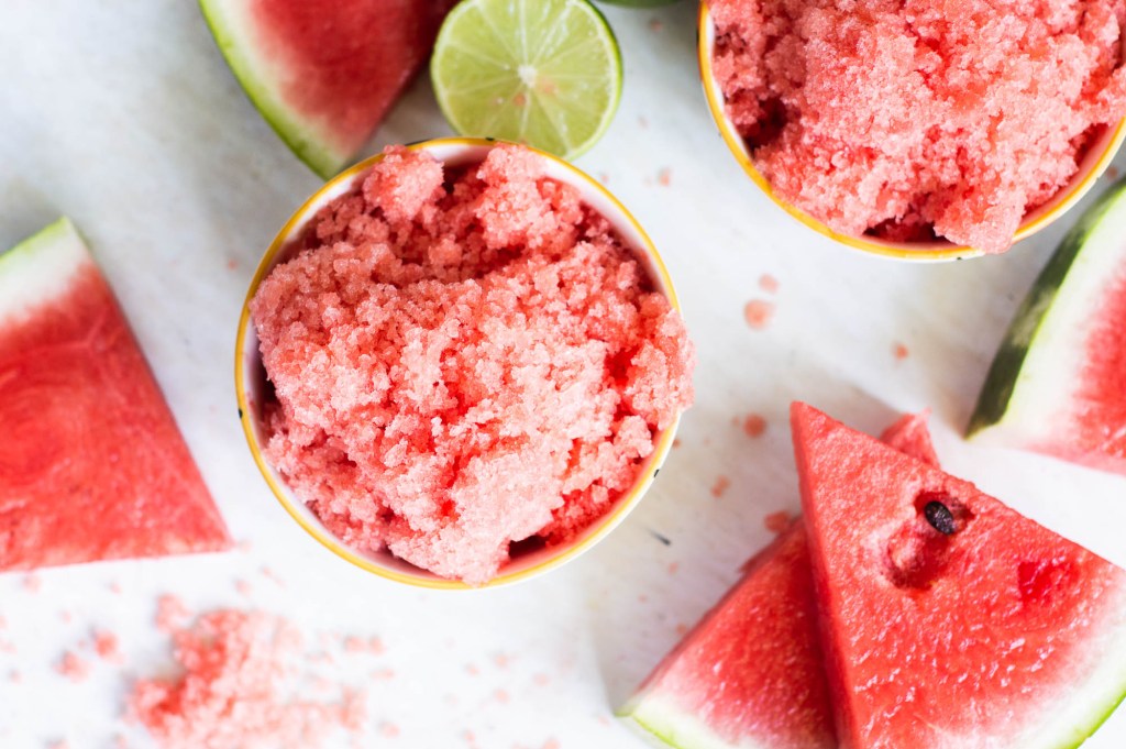 Overhead flat-lay of watermelon chunks, halved limes, fresh mint, sugar, and a baking dish on a marble surface — granita ingredients prep shot