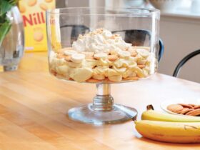 A large glass trifle dish filled with layered banana pudding, showing distinct layers of vanilla wafers, sliced bananas, and creamy vanilla pudding, topped with swirls of whipped cream and crushed vanilla wafers, shot in warm natural light on a rustic wooden table