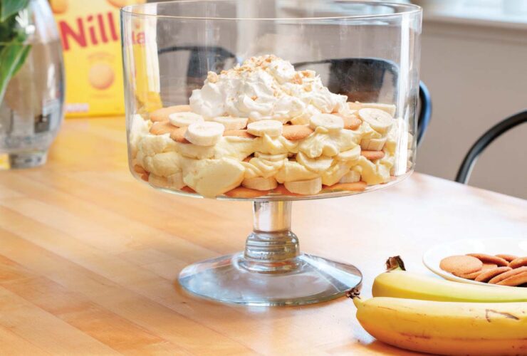 A large glass trifle dish filled with layered banana pudding, showing distinct layers of vanilla wafers, sliced bananas, and creamy vanilla pudding, topped with swirls of whipped cream and crushed vanilla wafers, shot in warm natural light on a rustic wooden table
