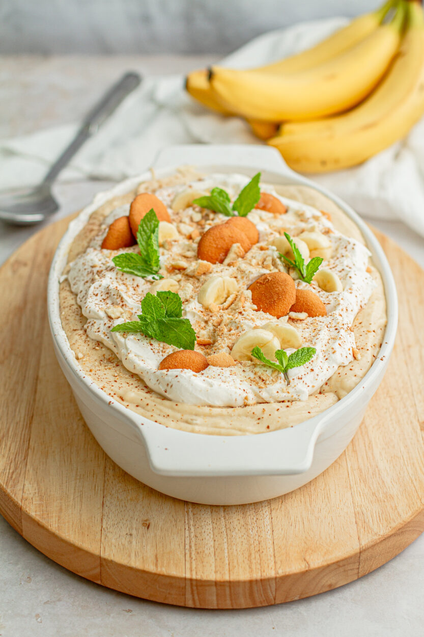 Overhead flat-lay of banana pudding ingredients including ripe bananas, a box of Nilla wafers, a block of cream cheese, cartons of heavy cream and whole milk, vanilla extract, sugar, cornstarch, and egg yolks, arranged on a white marble surface