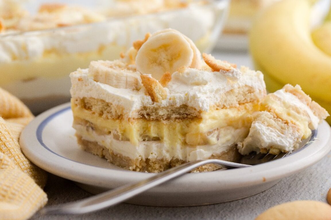 A close-up spoonful of banana pudding being lifted from a glass dish, showing distinct layers of creamy vanilla pudding, a softened vanilla wafer, and a slice of ripe banana, with whipped cream visible in the background, shot with shallow depth of field