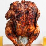 Beer Can Chicken finished dish — whole golden crispy grilled chicken standing upright on a beer can, plated on a wooden cutting board with fresh herbs and lemon slices
