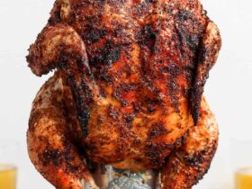 Beer Can Chicken finished dish — whole golden crispy grilled chicken standing upright on a beer can, plated on a wooden cutting board with fresh herbs and lemon slices
