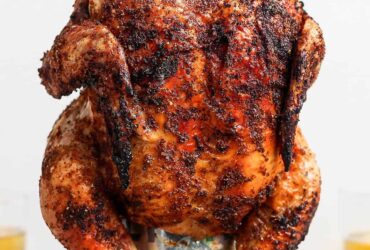 Beer Can Chicken finished dish — whole golden crispy grilled chicken standing upright on a beer can, plated on a wooden cutting board with fresh herbs and lemon slices