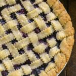 Golden lattice-top blueberry pie cooling on a wire rack, deep purple filling bubbling through the woven crust, dusted with coarse sugar, summer sunlight overhead