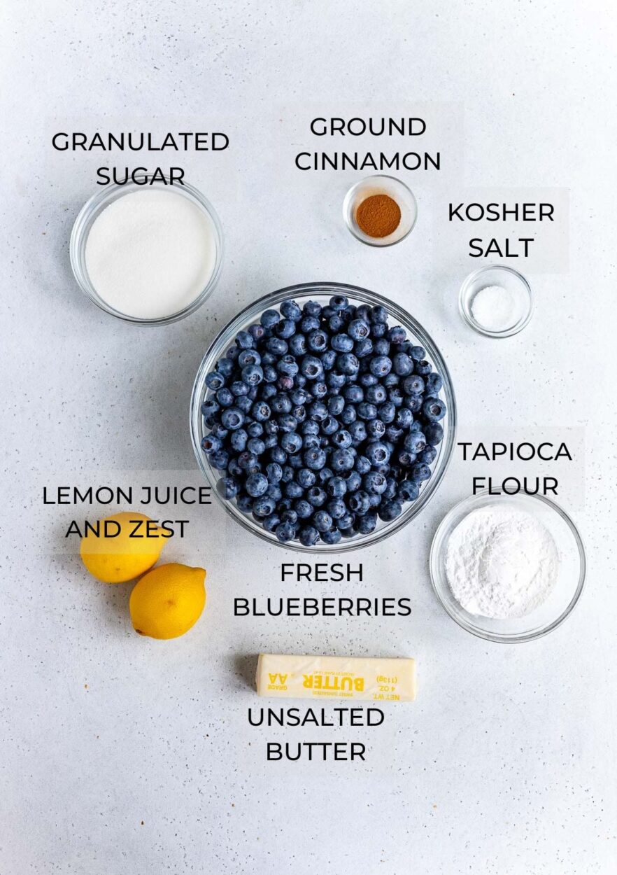 Overhead flat-lay of fresh blueberries, cold cubed butter, lemon, tapioca starch, sugar, and pie dough discs on a marble surface