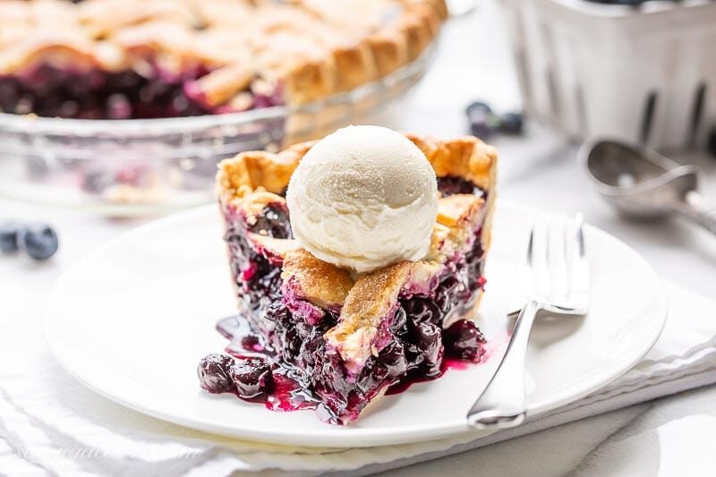 Close-up of a freshly cut slice of blueberry pie on a white plate, glossy deep-purple filling visible, flaky golden lattice crust, scoop of melting vanilla ice cream alongside