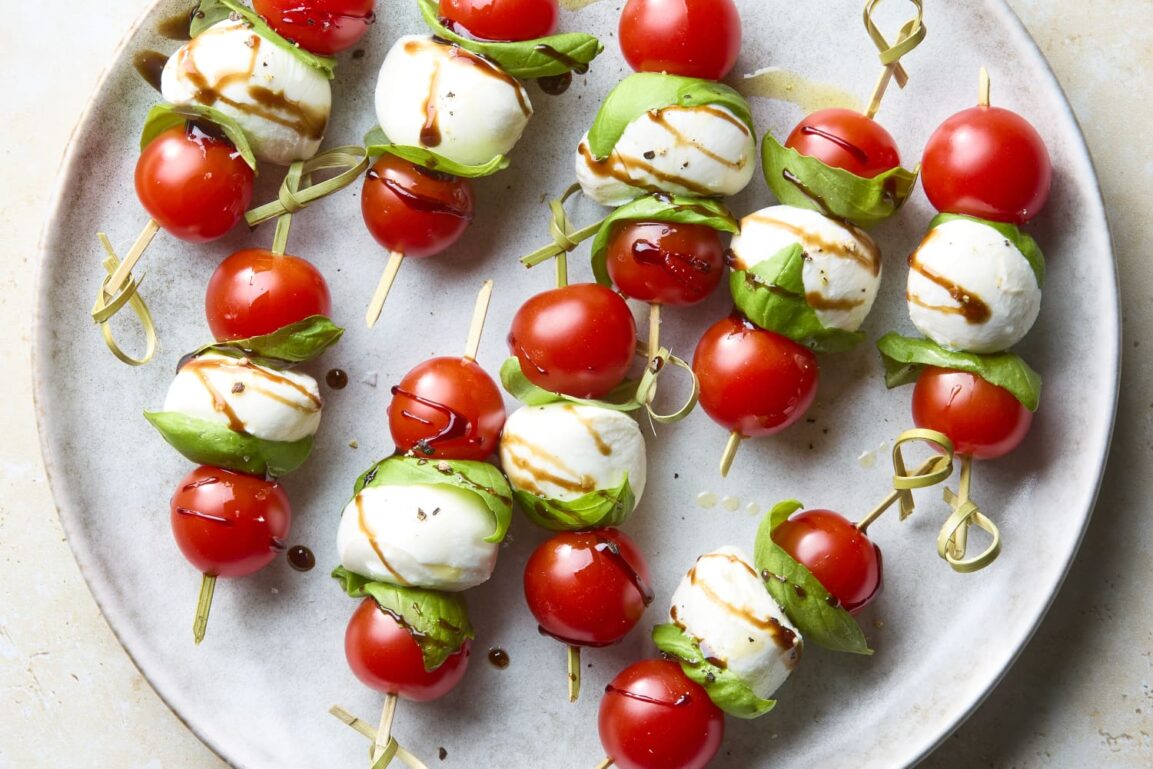 Caprese skewers with cherry tomatoes, fresh mozzarella, and basil on a white marble board drizzled with balsamic glaze