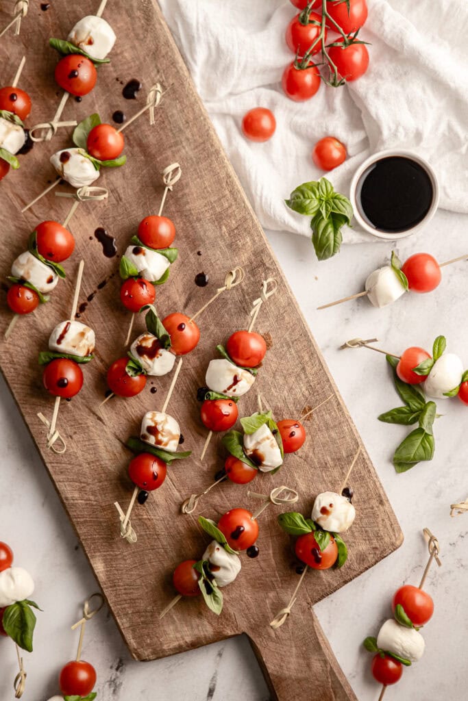 Caprese skewer ingredients laid out flat on a wooden board — cherry tomatoes, mozzarella pearls, fresh basil, olive oil, and balsamic glaze