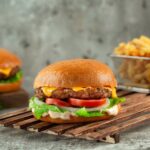 Classic Cheeseburgers stacked high with melted American cheese, lettuce, tomato, and pickles on a toasted brioche bun, served on a wooden board with fries in the background