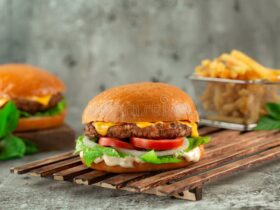Classic Cheeseburgers stacked high with melted American cheese, lettuce, tomato, and pickles on a toasted brioche bun, served on a wooden board with fries in the background