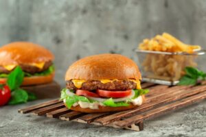 Classic Cheeseburgers stacked high with melted American cheese, lettuce, tomato, and pickles on a toasted brioche bun, served on a wooden board with fries in the background