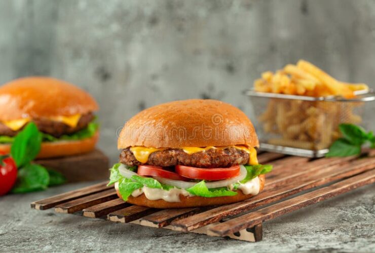 Classic Cheeseburgers stacked high with melted American cheese, lettuce, tomato, and pickles on a toasted brioche bun, served on a wooden board with fries in the background