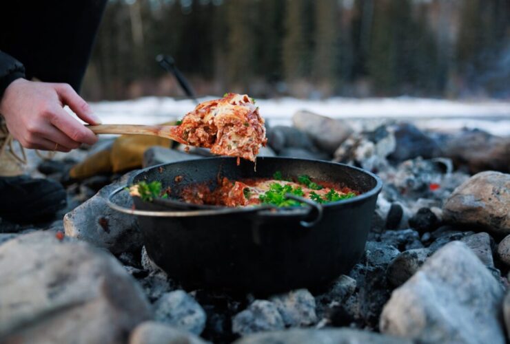 Dutch oven lasagna with bubbling cheese and golden top served at a campsite with pine trees in the background