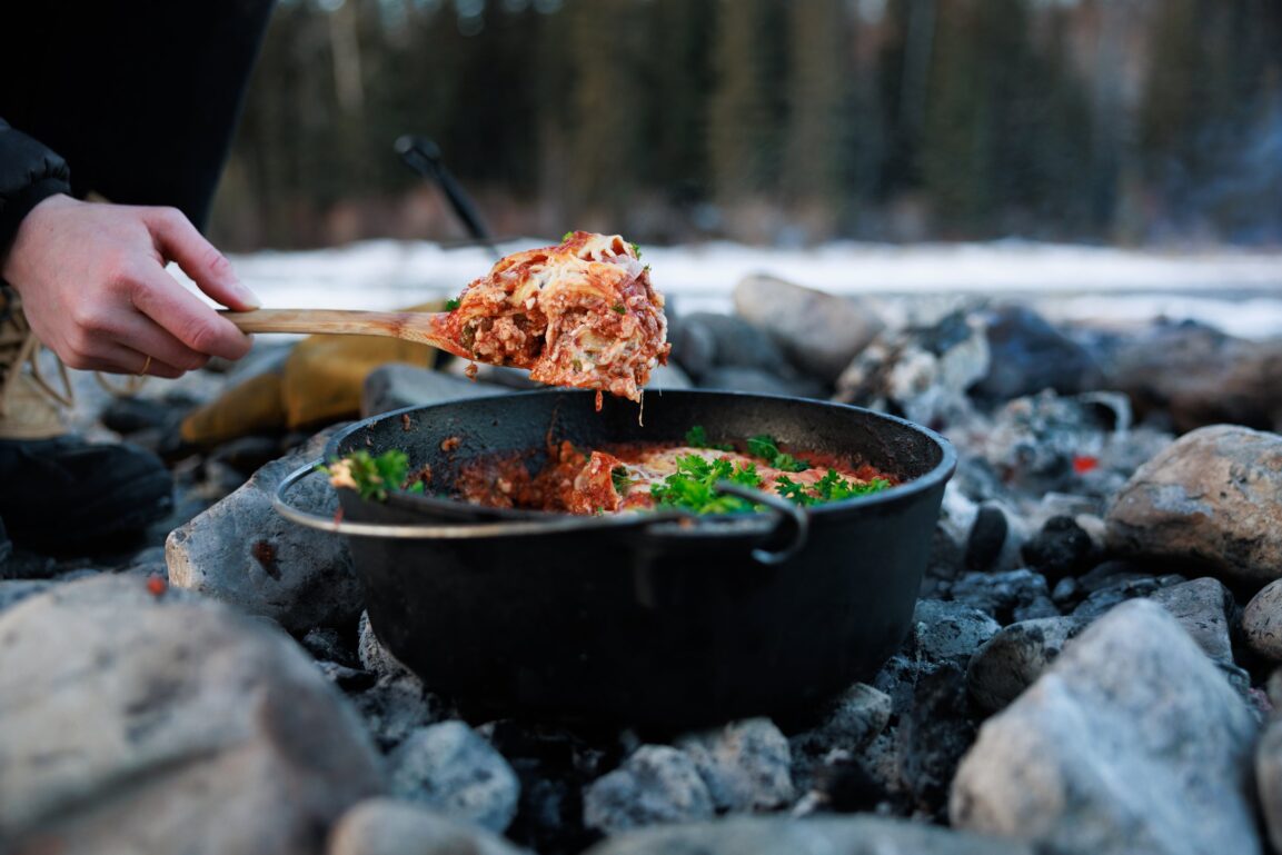 Dutch oven lasagna with bubbling cheese and golden top served at a campsite with pine trees in the background