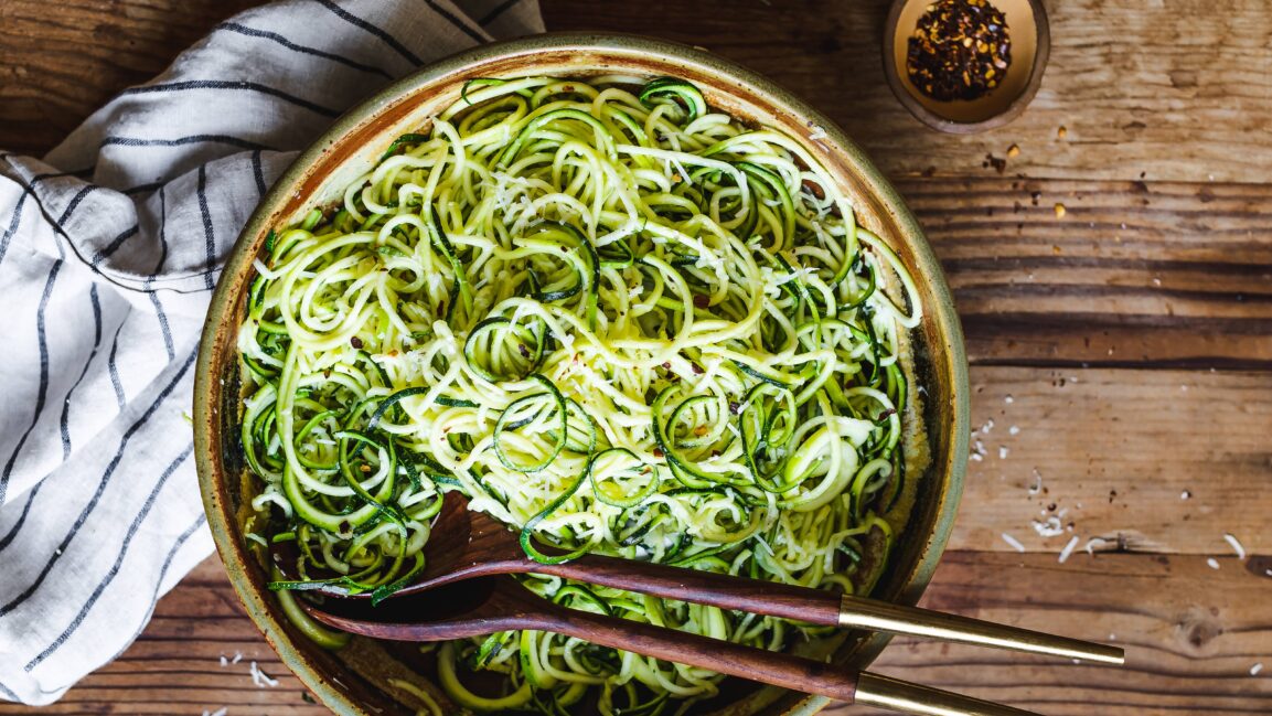 Overhead flat-lay of ingredients for Garlic Parmesan Zoodles — fresh zucchini, garlic cloves, Parmesan wedge, butter, olive oil, lemon, white wine, and fresh herbs on a marble surface