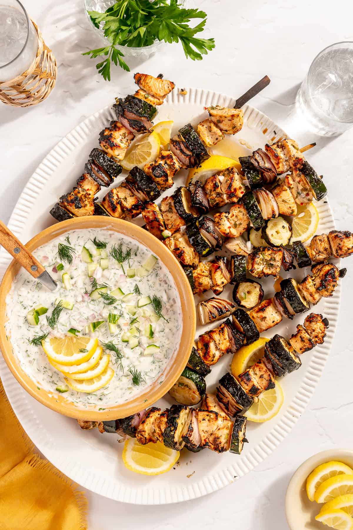 Close-up of grilled chicken skewer being dipped into thick, creamy tzatziki sauce with visible fresh dill, cucumber, and grill char marks on the chicken