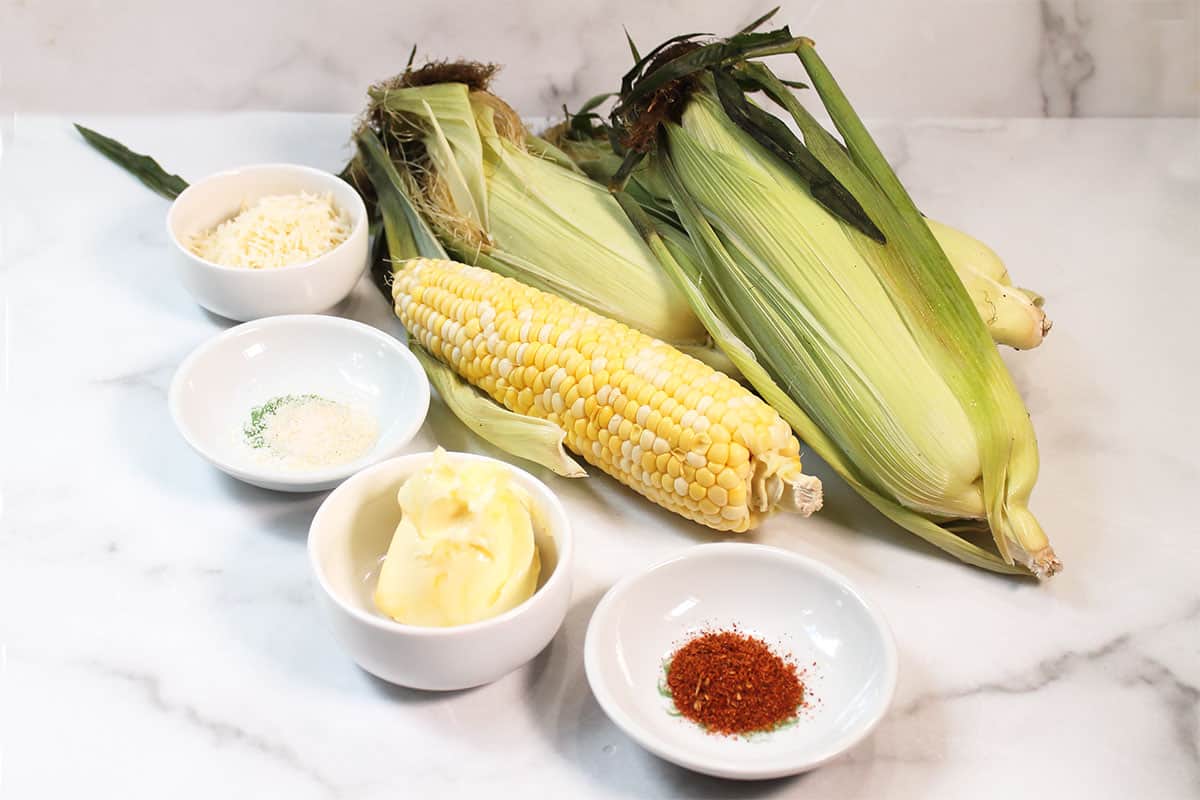 Ingredients for elote-style grilled corn laid out flat — ears of corn, butter, Parmesan, chili powder, lime, and mayonnaise on a marble surface