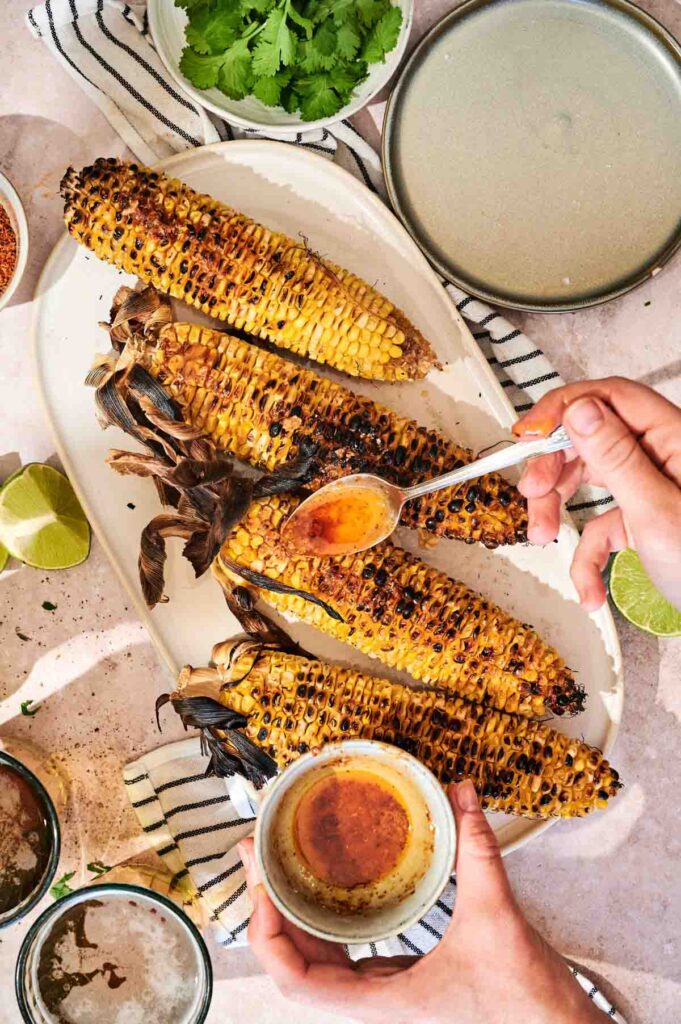 Close-up of elote-style grilled corn on the cob with melted Parmesan, chili powder, lime wedge, and fresh cilantro garnish