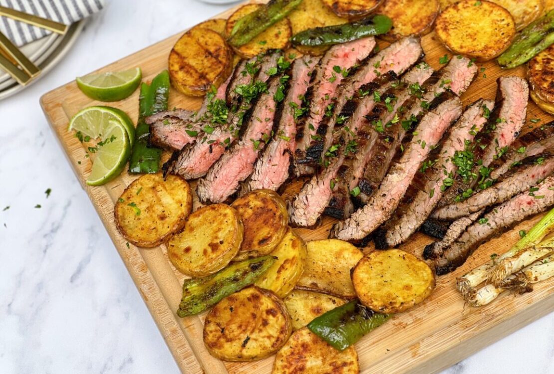 Grilled flank steak sliced thin on a wooden board surrounded by warm tortillas, fresh cilantro, lime wedges, and charred peppers — overhead beauty shot