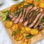 Grilled flank steak sliced thin on a wooden board surrounded by warm tortillas, fresh cilantro, lime wedges, and charred peppers — overhead beauty shot