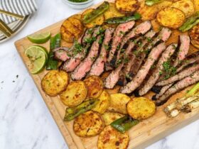 Grilled flank steak sliced thin on a wooden board surrounded by warm tortillas, fresh cilantro, lime wedges, and charred peppers — overhead beauty shot