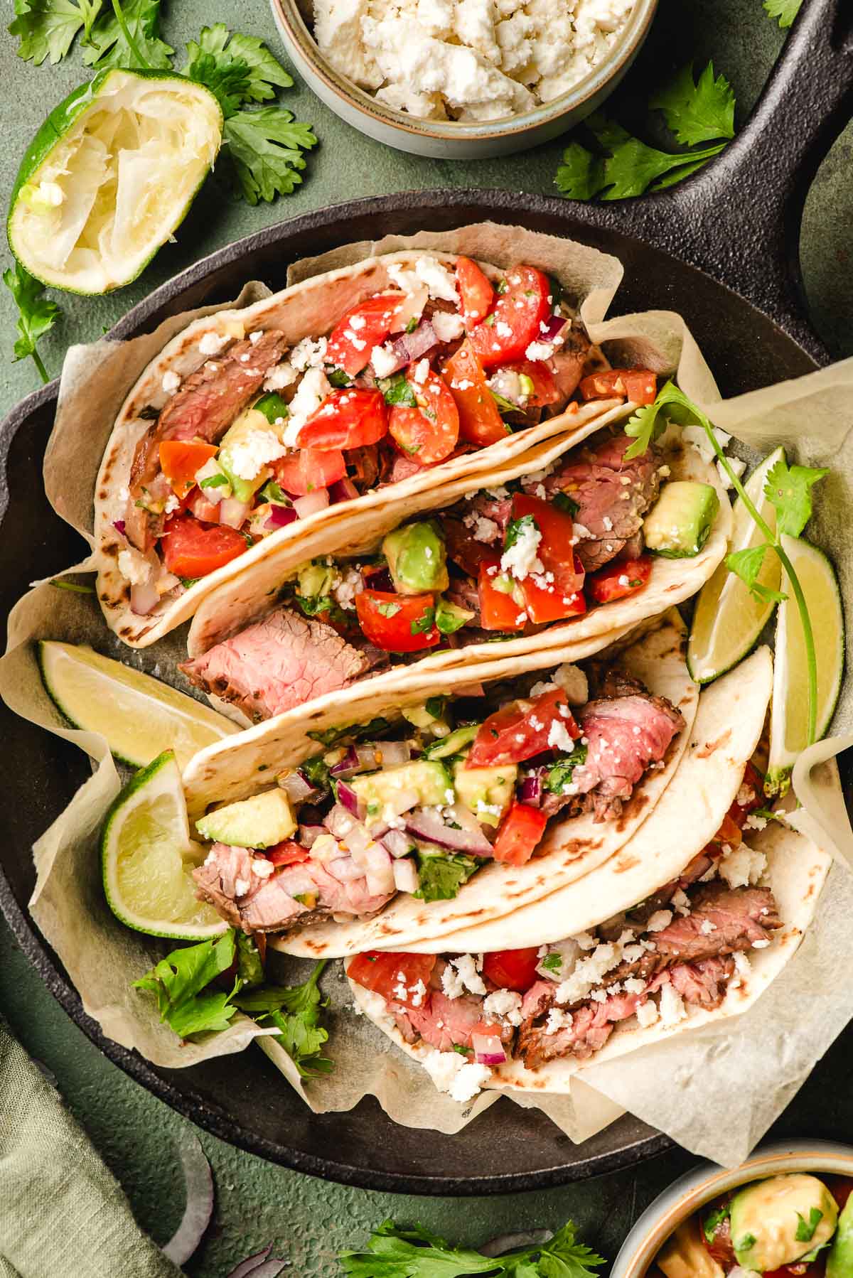 Close-up of two grilled flank steak tacos in charred corn tortillas topped with pico de gallo, avocado, pickled onions, and fresh cilantro with lime wedges on the side
