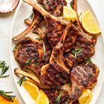 Grilled lamb chops plated on a white platter with fresh rosemary sprigs, lemon wedges, and a golden garlic crust, shot from above on a rustic wooden table
