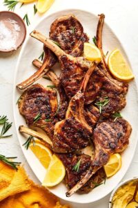 Grilled lamb chops plated on a white platter with fresh rosemary sprigs, lemon wedges, and a golden garlic crust, shot from above on a rustic wooden table