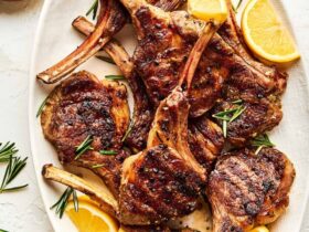 Grilled lamb chops plated on a white platter with fresh rosemary sprigs, lemon wedges, and a golden garlic crust, shot from above on a rustic wooden table