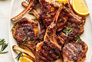 Grilled lamb chops plated on a white platter with fresh rosemary sprigs, lemon wedges, and a golden garlic crust, shot from above on a rustic wooden table