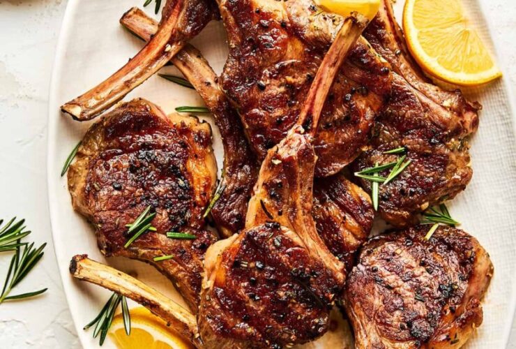 Grilled lamb chops plated on a white platter with fresh rosemary sprigs, lemon wedges, and a golden garlic crust, shot from above on a rustic wooden table