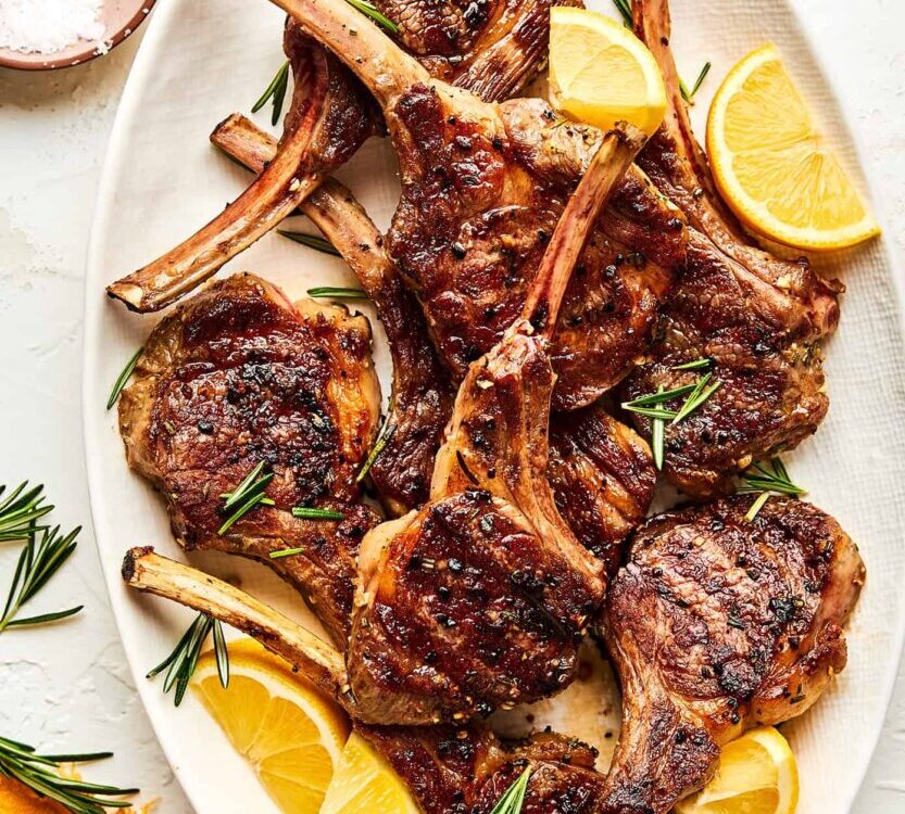 Grilled lamb chops plated on a white platter with fresh rosemary sprigs, lemon wedges, and a golden garlic crust, shot from above on a rustic wooden table