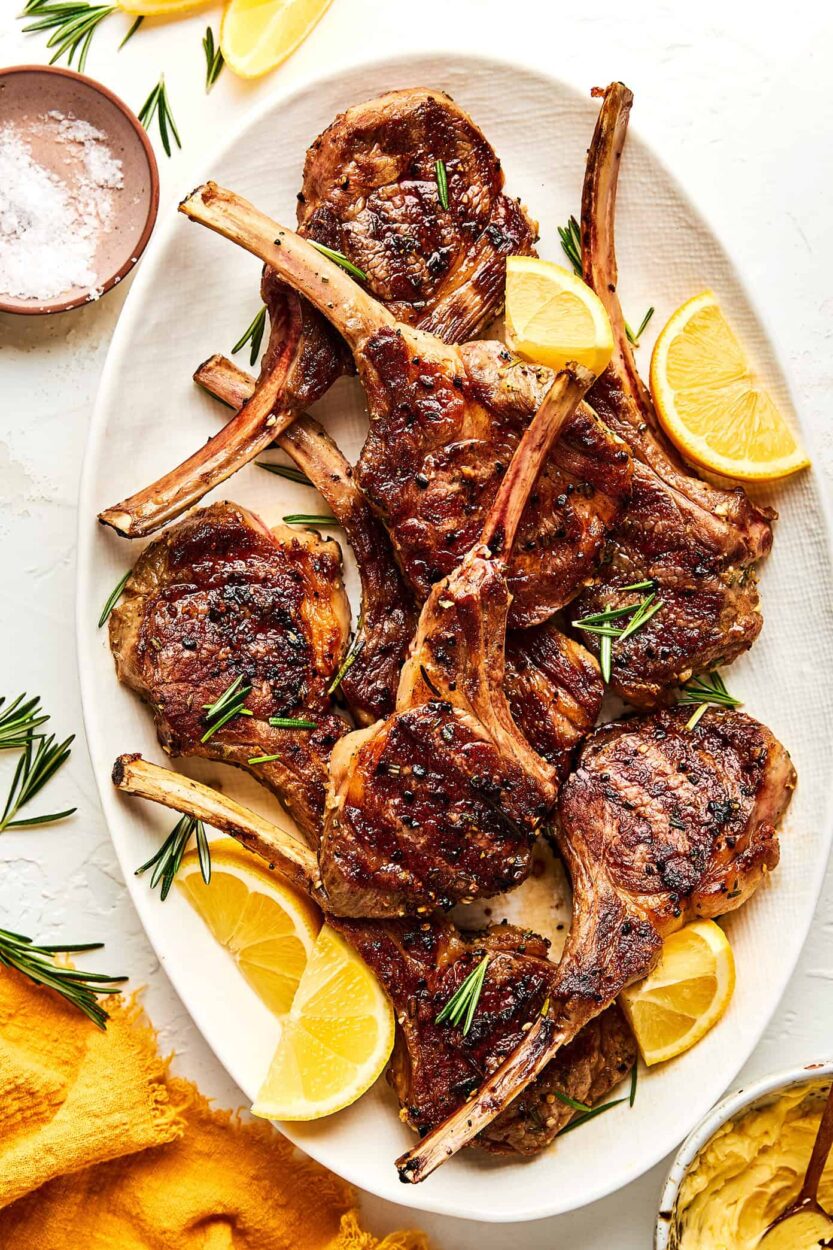 Grilled lamb chops plated on a white platter with fresh rosemary sprigs, lemon wedges, and a golden garlic crust, shot from above on a rustic wooden table