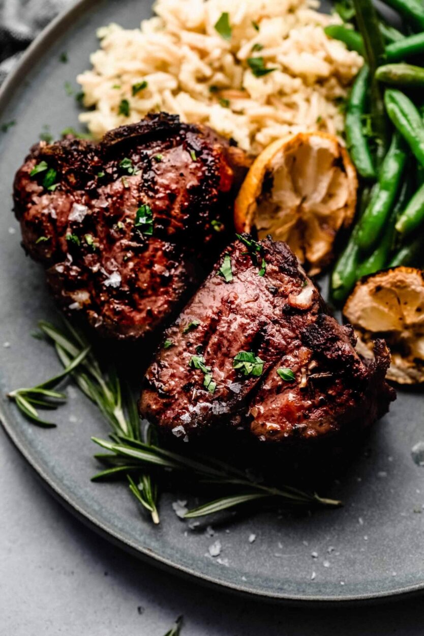 Lamb chops in a glass dish submerged in a rosemary garlic olive oil marinade with lemon zest and cracked black pepper, overhead shot on a marble surface