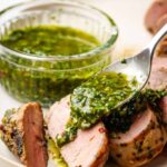 Grilled pork tenderloin sliced on a wooden cutting board with fresh herbs, roasted garlic, and a side of chimichurri sauce