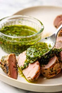Grilled pork tenderloin sliced on a wooden cutting board with fresh herbs, roasted garlic, and a side of chimichurri sauce
