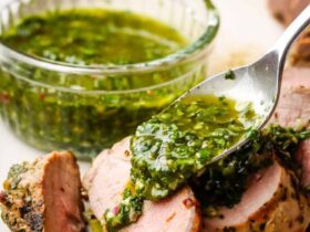 Grilled pork tenderloin sliced on a wooden cutting board with fresh herbs, roasted garlic, and a side of chimichurri sauce