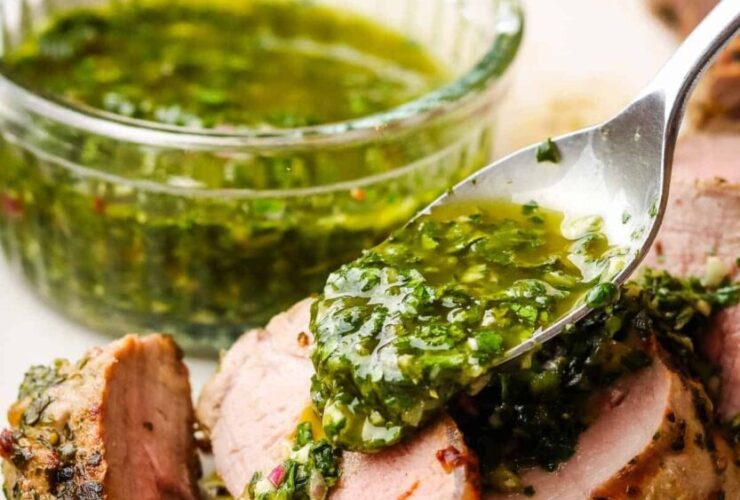 Grilled pork tenderloin sliced on a wooden cutting board with fresh herbs, roasted garlic, and a side of chimichurri sauce