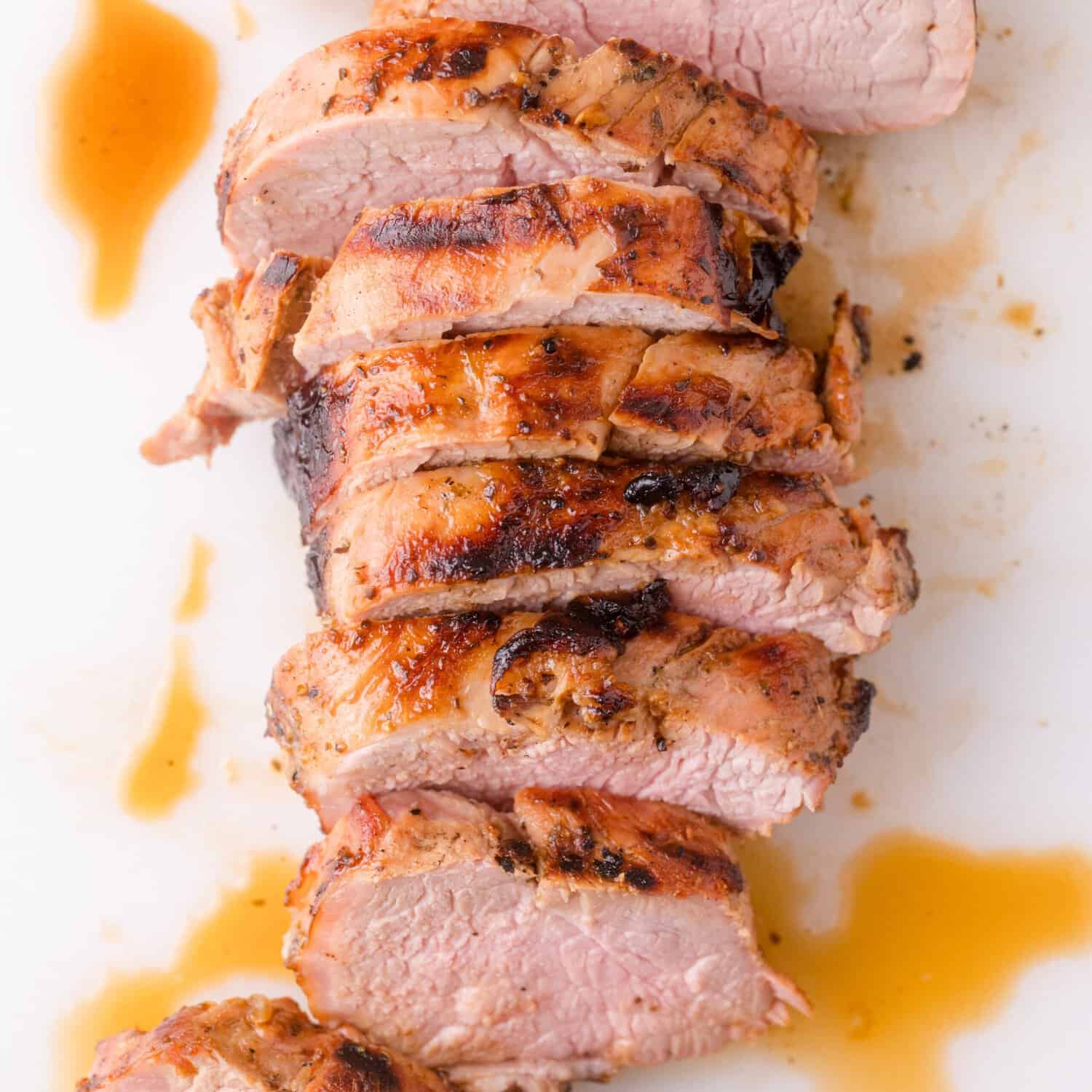 Close-up of grilled pork tenderloin slices fanned out on a white platter, showing the blush-pink interior, charred crust, and garnished with fresh rosemary and a drizzle of chimichurri